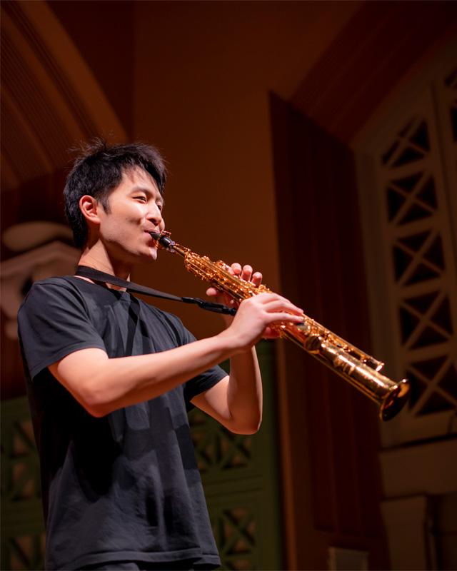 A male Asian student playing the telesaxophone, in a dark performance space.